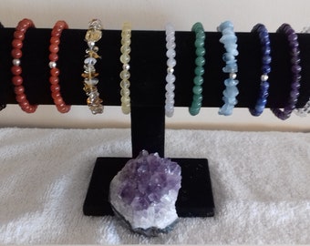Könnte beinhalten: Eine Auswahl von zehn Armbändern aus verschiedenen Edelsteinen, darunter schwarzer Onyx, roter Karneol, Bernstein, gelber Jaspis, Rosenquarz, grüner Aventurin, Aquamarin, Lapislazuli, Amethyst und klarer Quarz. Die Armbänder sind auf einem schwarzen Samtständer mit einem großen Amethystkristall an der Basis angeordnet.