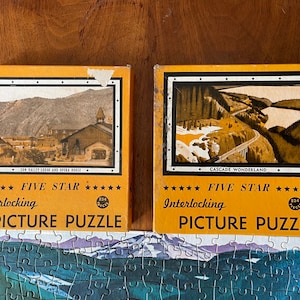 May include: Two vintage Five Star Interlocking Picture Puzzle boxes. The boxes are yellow with black text and a picture of a mountain scene. The text on the boxes reads "Sun Valley Lodge and Opera House" and "Cascade Wonderland".