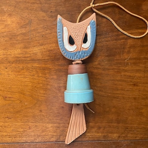 May include: A ceramic owl wind chime with a blue and brown painted design. The owl has large, black eyes and a brown body with blue accents. The chime is suspended from a brown cord.