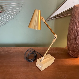 May include: A gold desk lamp with a flexible arm and a white base. The lamp is on a wooden surface.