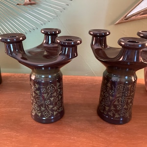 May include: Two dark brown ceramic candle holders. Each has three candle holders and a gold floral pattern. The candle holders are on a wooden surface.