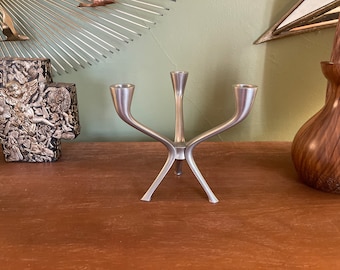 May include: A silver three-armed candle holder with a modern design. The candle holder is made of metal and has a sleek, minimalist look.