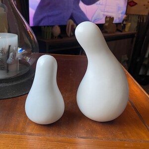 以下が含まれることがあります： 木製の表面に置かれた、白い陶器製の梨の形をした彫刻が2つ。大きな梨は小さな梨よりも背が高く、幅広いです。