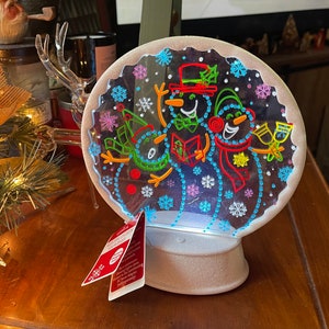 Könnte beinhalten: Eine weiße Schneekugel mit einer durchsichtigen Kuppel, die drei Schneemänner mit bunten Umrissen zeigt. Die Schneemänner singen Weihnachtslieder und halten ein Buch und ein Glas Champagner. Die Schneekugel ist von innen beleuchtet und hat einen weißen Sockel.