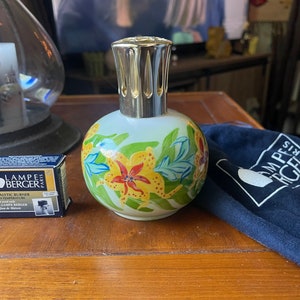 May include: A white ceramic oil diffuser with a gold metal lid and a floral design. The diffuser is sitting on a wooden table with a small box that says "Lampe Berger Paris" and a blue bag with the same text.