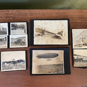 May include: A vintage photo album filled with black and white photographs of early aeroplanes. The album includes photos of biplanes, a blimp, and people standing near the aircraft.