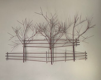 Pode incluir: Uma escultura de parede de metal que representa três árvores com ramos e uma cerca. A escultura é feita de metal fino marrom e está montada em uma parede branca.