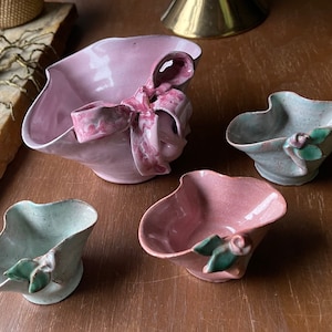 May include: Four ceramic bowls with decorative floral and ribbon accents. The bowls are in shades of pink and green.