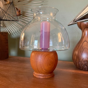 Può includere: Un portacandele in legno con una cupola di vetro e una candela viola all'interno. Il portacandele ha una base rotonda e una finitura liscia e lucida.