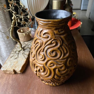 Puede incluir: Un jarrón de cerámica marrón con un patrón de espiral. El jarrón tiene un borde marrón oscuro y un acabado brillante.