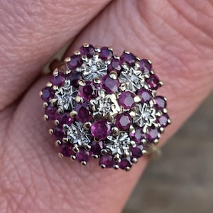 Könnte beinhalten: Ein goldener Ring mit einem Cluster aus tiefroten Edelsteinen und kleineren, klaren Steinen. Das Design des Rings ist ein Blumen- oder Sternmuster, wobei die Edelsteine eng beieinander liegen.