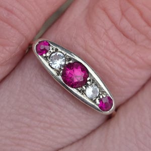 May include: A silver ring with an elongated oval design. The ring features a central, large, oval-cut, magenta gemstone, flanked by two smaller, round, white gemstones and two magenta gemstones.