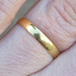 May include: Close-up of a gold ring. The ring is a solid band with a smooth, polished surface. The gold color is bright and reflective, with slight variations in tone. The ring is worn on a finger.