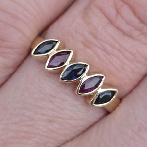 May include: A gold ring featuring five marquise-cut gemstones arranged in a row. The stones alternate between dark blue sapphires and deep red rubies, set in a polished gold band that encircles the finger with a sleek, multi-stone design.