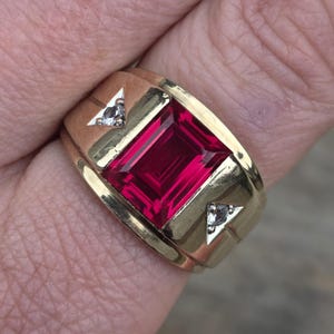 Puede incluir: Un anillo de oro con una gran piedra preciosa rectangular de color rojo intenso. El anillo tiene una banda gruesa y está acentuado con pequeñas incrustaciones triangulares que contienen piedras transparentes. El anillo es un diseño clásico.