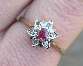 Pode incluir: Um anel de ouro com um design em forma de flor. O centro da flor é uma pedra preciosa redonda, vermelha escura, rodeada por pedras preciosas claras menores. A banda do anel é uma banda de ouro fina e simples.