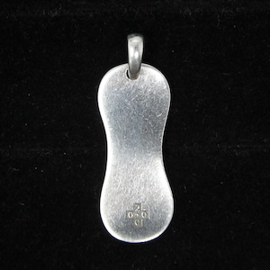May include: A silver-toned metal pendant with a smooth, elongated, hourglass-like shape and a rounded bail at the top. The surface features a brushed texture and a small, stamped cross-shaped hallmark near the bottom containing the numbers 2, 0, 1, and 0.