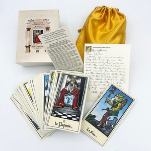 Puede incluir: Una baraja de cartas del Tarot con una caja dorada y blanca, una bolsa de satén amarilla y una nota manuscrita. Las cartas presentan ilustraciones coloridas en estilo renacentista. La nota dice "Querido/a [Nombre], Hoy recibes un nuevo juego de trabajo maravilloso que encarna tanto las ideas tradicionales sobre los misterios del camino como los pasos del tarot. Que la luz del corazón del amor guíe el viaje del Ser. Estoy seguro de que este conjunto de tarot te guiará hacia las alturas y las profundidades de esta vida.  Bill esto [date]"