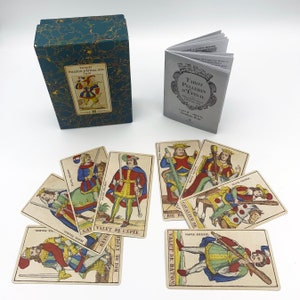Puede incluir: Un juego de cartas de tarot de la baraja Pellerin d'Epinal. Las cartas presentan ilustraciones coloridas de personas y símbolos. La caja es azul verdoso con detalles dorados. El folleto es blanco con texto negro. Las cartas están extendidas sobre una superficie blanca.
