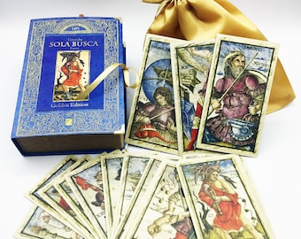 Könnte beinhalten: Eine blaue Schachtel mit goldenen Akzenten und dem Text "Sola Busca Tarot Golden Edition" auf der Vorderseite. Die Schachtel enthält ein Tarot-Kartendeck mit Illustrationen von Personen und Szenen. Die Karten liegen auf einer weißen Oberfläche ausgebreitet, und im Hintergrund befindet sich eine goldene Satintüte.