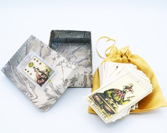May include: An open box and a deck of tarot cards with a gold satin drawstring bag. The box has a marbled design and a small card with text and an illustration. The cards are fanned out, and one card is visible with the text "LE BATELEUR".
