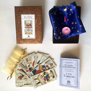 Pode incluir: Um conjunto de cartas de tarot com uma caixa castanha, uma bolsa de cetim azul com estrelas douradas, uma vela rosa e um pequeno livro. As cartas de tarot estão em leque, exibindo ilustrações coloridas.