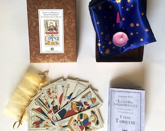 May include: A tarot card set featuring a brown box, a blue satin pouch with gold stars, a pink candle, and a small book. The tarot cards are fanned out, showing colourful illustrations.