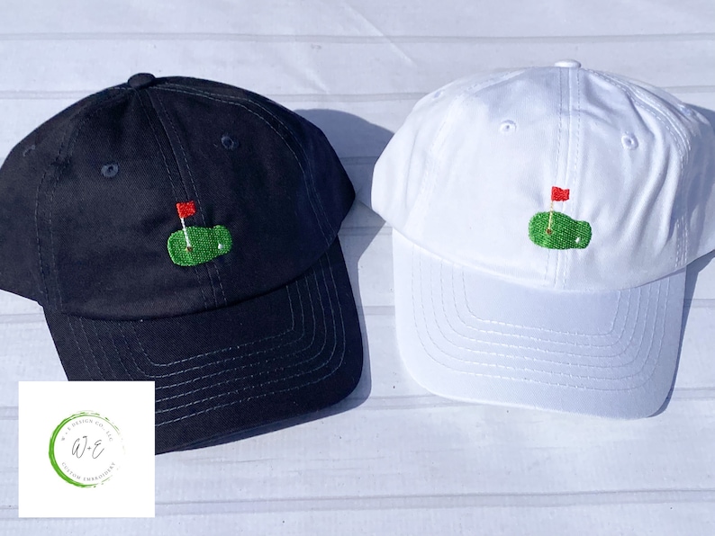 Op de afbeelding: Twee baseballpetten met een geborduurd groen golfveld en een rode vlag. De ene pet is zwart en de andere is wit.