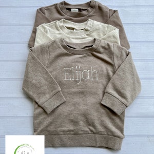 May include: Three brown, beige, and cream colored sweatshirts stacked on top of each other. The bottom sweatshirt has the name "Elijah" embroidered on the front in white thread.