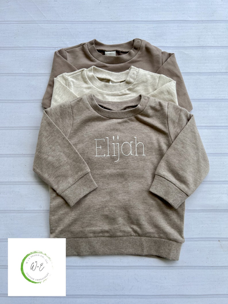 May include: Three brown, beige, and cream colored sweatshirts stacked on top of each other. The bottom sweatshirt has the name "Elijah" embroidered on the front in white thread.
