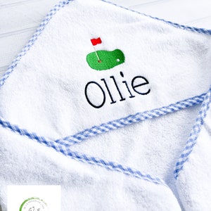 May include: White hooded towel with blue and white gingham trim. The towel has an embroidered green golf course with a red flag and the name "Ollie" in black.