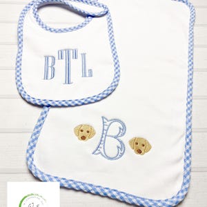 May include: A set of two white baby bibs with blue and white gingham trim. One bib has the monogram "BTL" embroidered on it. The other bib has a large embroidered letter "B" and two embroidered yellow labrador puppy faces.
