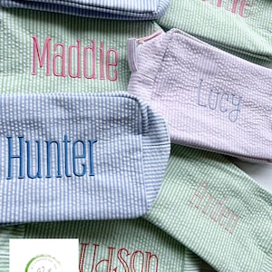 May include: A collection of personalized green, blue, and pink striped fabric bags with names embroidered on them. The names include Maddie, Hunter, Lucy, Annie, and Upson.