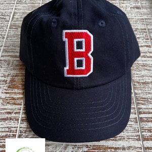 May include: A navy blue baseball cap with a red embroidered letter "B" on the front. The cap has a curved brim and white stitching detail around the letter.