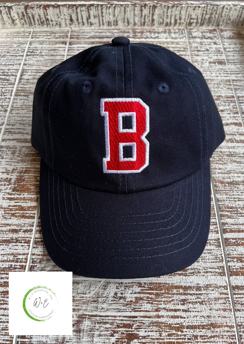 May include: A navy blue baseball cap with a red embroidered letter "B" on the front. The cap has a curved brim and white stitching detail around the letter.