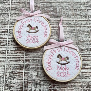 Pode incluir: Dois enfeites de Natal personalizados para o primeiro Natal de um beb&ecirc;, de cor rosa e branca, com um design de cavalo de balan&ccedil;o. Os enfeites s&atilde;o bordados com os nomes "Aida" e "Molly" e o ano "2024".