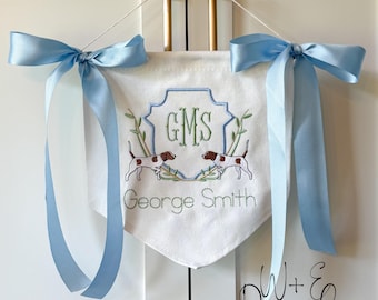 May include: White banner with light blue accents, featuring embroidered details. The banner has a light blue ribbon tied in a bow on each side. The banner has the initials "GMS" and the name "George Smith" embroidered on it, along with two dog illustrations.