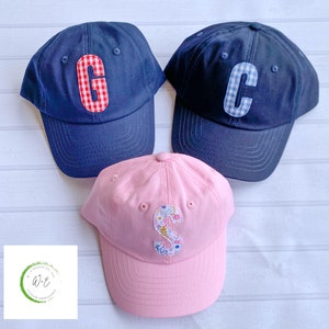 May include: Three personalized baseball caps. One is navy blue with a red and white gingham letter C. Another is navy blue with a blue and white gingham letter C. The third is pink with a floral letter S.