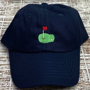 Op de afbeelding: Een donkerblauwe baseballpet met een geborduurd groen golfveld en een rode vlag.