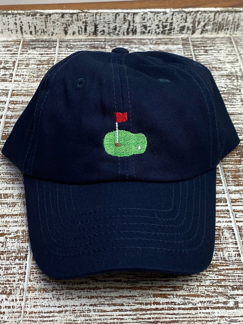 Op de afbeelding: Een donkerblauwe baseballpet met een geborduurd groen golfveld en een rode vlag.