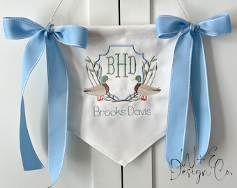 Op de afbeelding: Een witte stoffen banner met een monogram van de letters "BHD" in een groene krans met twee eenden aan weerszijden. De banner heeft ook de tekst "Brooks Davis" geborduurd onder de krans.