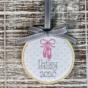 May include: A white fabric ornament with pink ballet shoes and the name "Hailey 2020" embroidered on it. The ornament is attached to a gray ribbon.