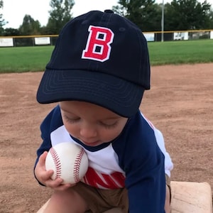 May include: A toddler wearing a navy blue baseball cap with a red and white letter "B" embroidered on the front. The child is holding a white baseball in their hand. The child is wearing a white and blue striped shirt with a red stripe.