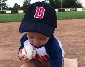 Op de afbeelding: Een peuter draagt een donkerblauwe baseballpet met een rode en witte letter "B" geborduurd op de voorkant. Het kind houdt een witte honkbal in zijn hand. Het kind draagt een wit en blauw gestreept shirt met een rode streep.