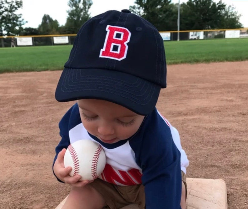 May include: A toddler wearing a navy blue baseball cap with a red and white letter "B" embroidered on the front. The child is holding a white baseball in their hand. The child is wearing a white and blue striped shirt with a red stripe.