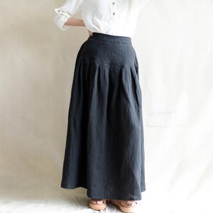Puede incluir: Una falda larga de color gris oscuro con cintura alta y pliegues. La falda está hecha de una tela fluida y llega hasta los tobillos. La modelo lleva una blusa blanca. La falda tiene un bolsillo en la parte delantera.