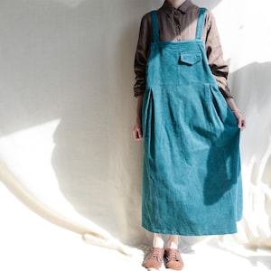 May include: A teal blue corduroy overall dress with thin straps. The dress has a single pocket on the front and a slightly flared skirt.