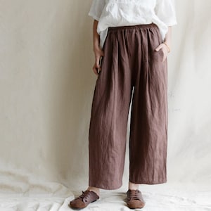 May include: Wide-leg brown linen pants with an elastic waistband and pockets. The pants are a solid color and appear to be made of a lightweight fabric. The model is wearing brown shoes and a white top.