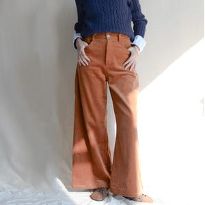 May include: Wide-leg, rust-colored corduroy pants. The pants have a high waist and pockets. The model is wearing a navy blue cable-knit sweater and a light blue and white striped shirt underneath.