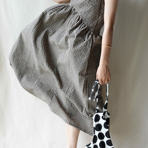 May include: A black and white gingham dress with a loose, flowing silhouette. The dress has a sleeveless design and a round neckline. The model is wearing brown leather shoes.